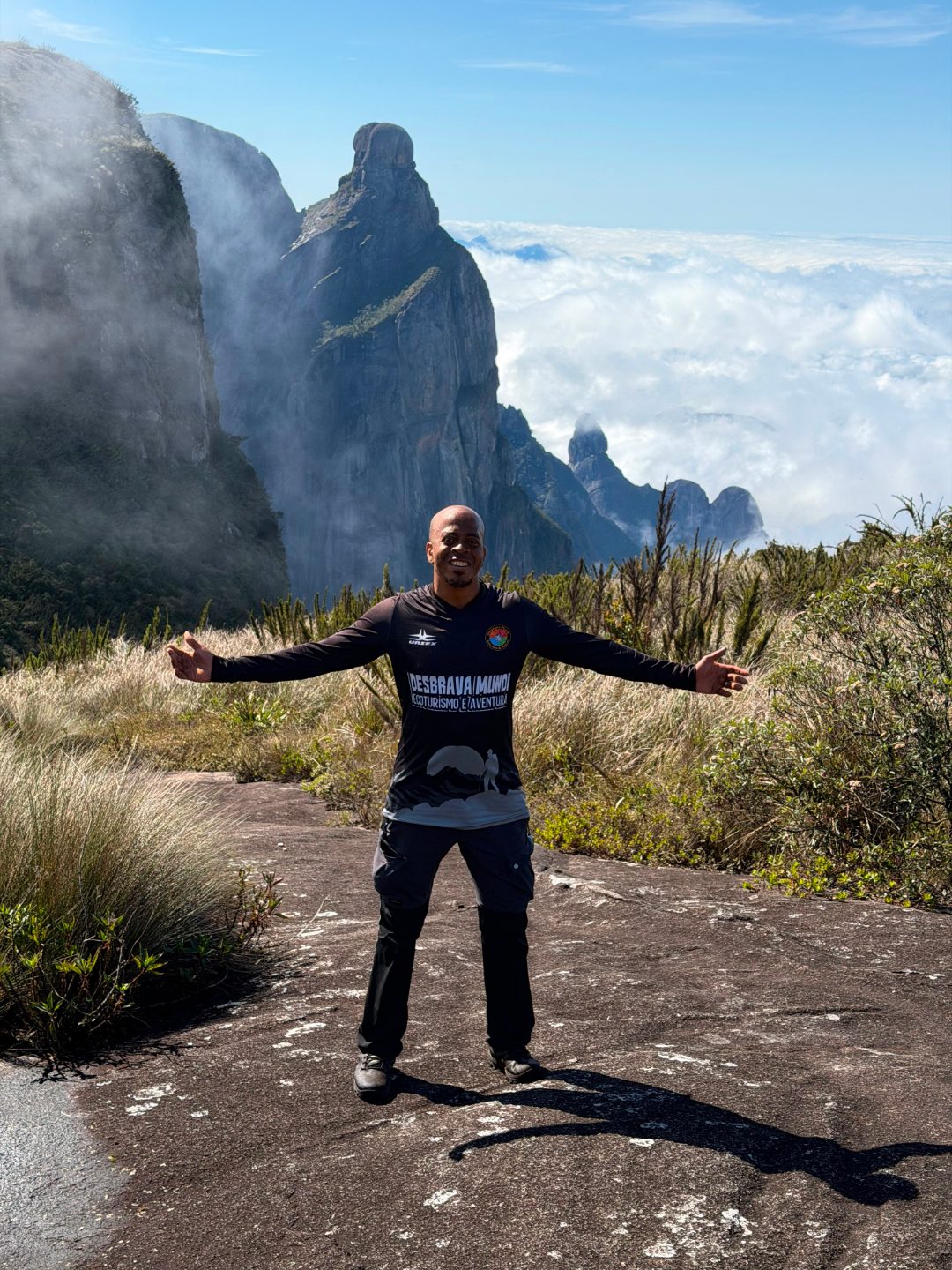 Ricardo de braços abertos no alto da serra, com picos de pedra e um mar de nuvens ao fundo